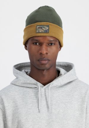 Green and yellow knit beanie with a patch, ribbed texture, and a fold-over design; worn with a light gray hooded sweatshirt.