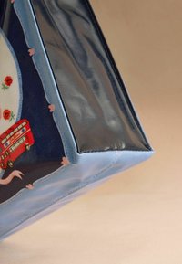 Blue tote bag corner with floral pattern and red double-decker bus design, featuring shiny textured side panel and stitched edges on beige background.