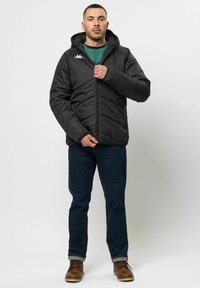 Veste noire matelassée avec capuche, présentant des coutures en chevrons et un logo blanc sur la poitrine gauche. Associée à un jean bleu foncé et des bottes marron.