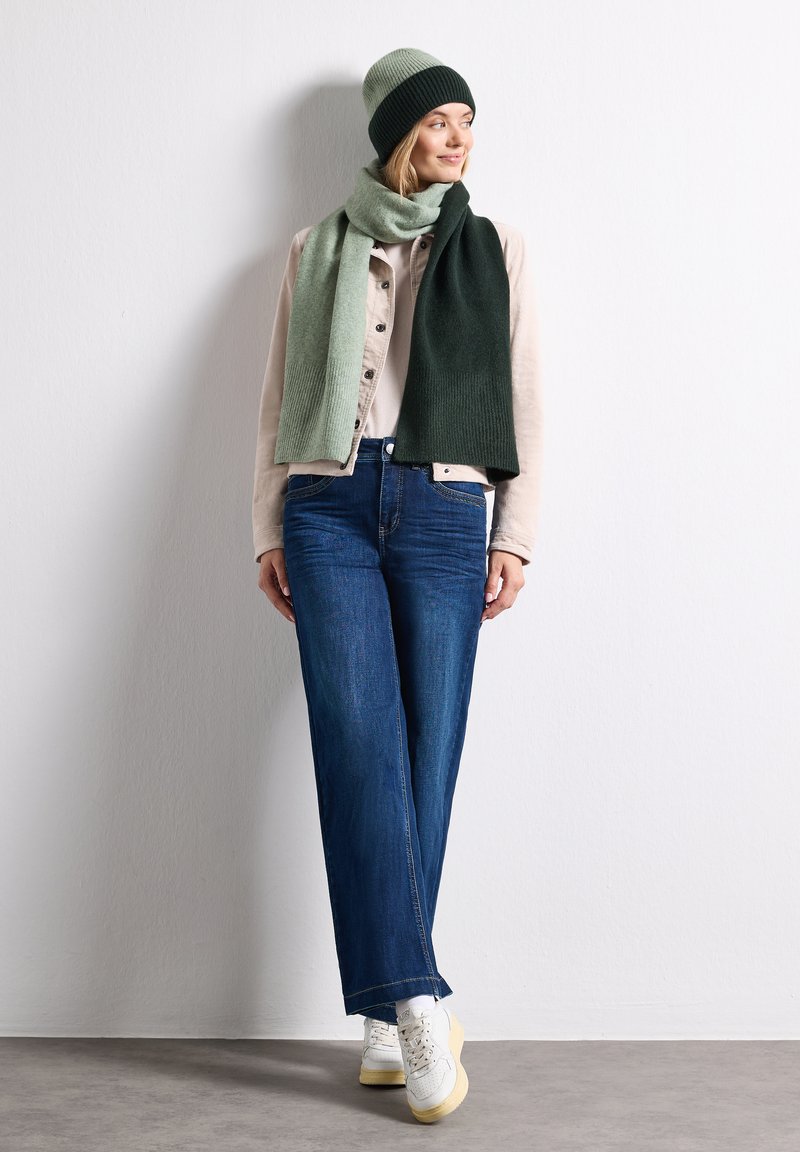 Femme portant un bonnet vert et une écharpe bicolore verte, une veste beige, un jean bleu et des baskets blanches, debout contre un mur blanc uni.
