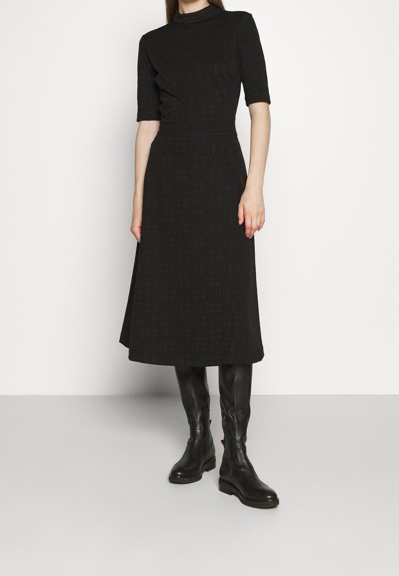 Femme portant une robe noire mi-longue avec des manches aux coudes et des bottes en cuir noires montant jusqu'aux genoux, debout sur un fond blanc.
