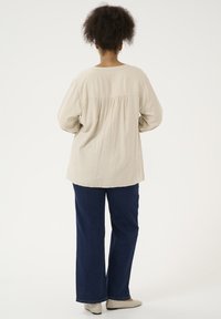 Beige Langarmoberteil mit gerafftem Rücken, kombiniert mit dunkelblauen weit geschnittenen Jeans und hellen Schuhen, vor einem weißen Hintergrund präsentiert.