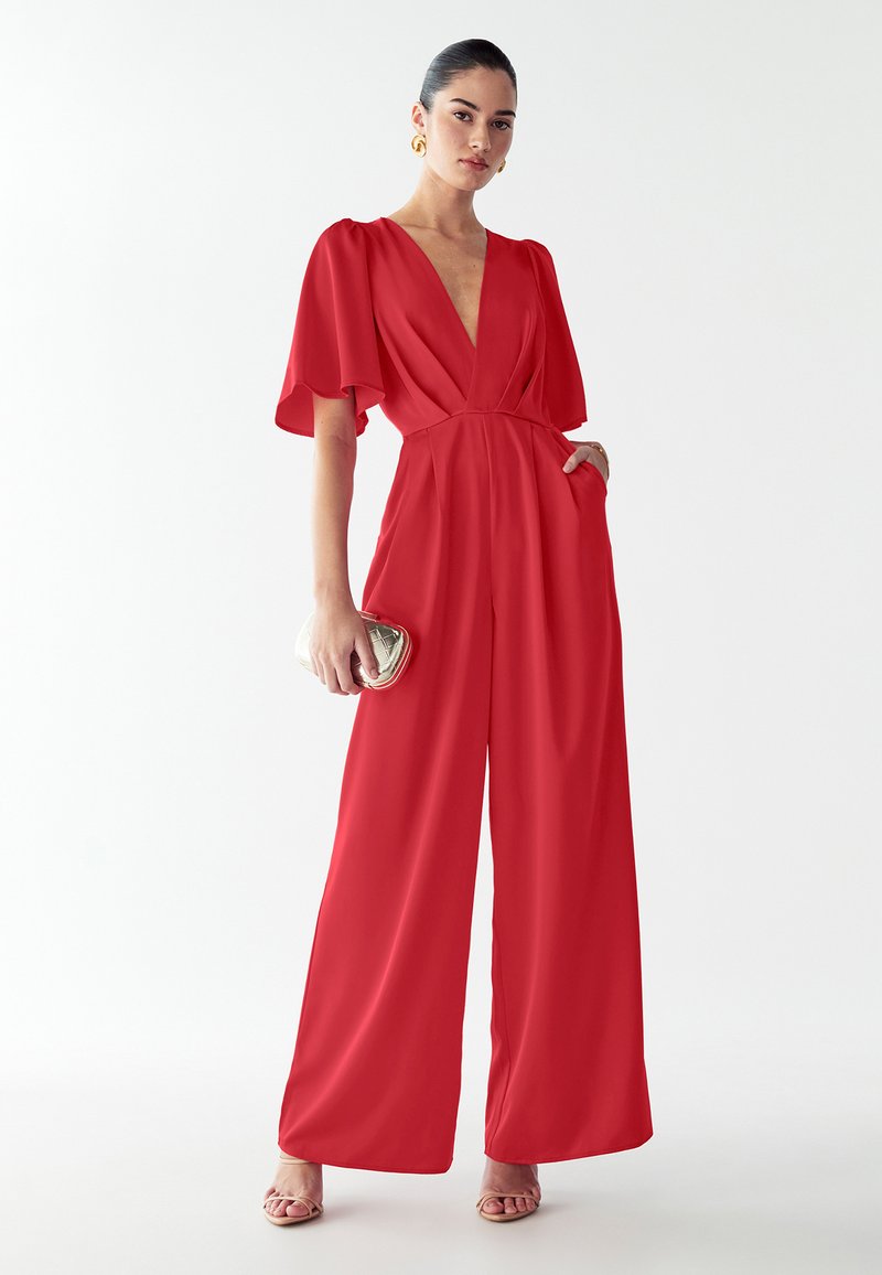 Red jumpsuit with short flutter sleeves, deep V-neck, and wide-leg design. Features side pockets and a smooth texture. Holding a small silver clutch.