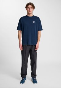 Marinblå bomullströja med rund halsringning och vitt chevronlogotyp på vänster bröst, kombinerad med svarta byxor och blåa träningsskor.