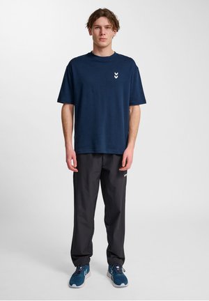 Marineblå bomulds T-shirt med rund halsudskæring og hvidt chevron-logo på venstre bryst, parret med sorte bukser og blå træningssko.