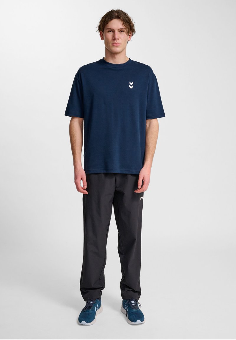 Marinblå bomullströja med rund halsringning och vitt chevronlogotyp på vänster bröst, kombinerad med svarta byxor och blåa träningsskor.