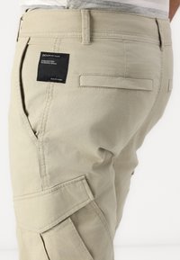 Svijetlo bež cargo hlače s bočnim džepovima, šivenim uzorkom i crnom etiketom na pojasu na kojoj piše "DENIM". Imaju glatku teksturu.