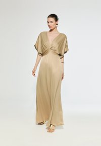 Brown satin maxi dress with short batwing sleeves and a knotted front detail. Accompanied by gold sandals and large circular earrings.
