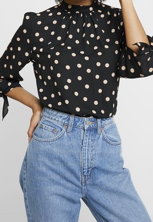 Mujer con blusa negra de manga larga con lunares beige y vaqueros de tiro alto azul claro, un brazo doblado y mano cerca de la cara.