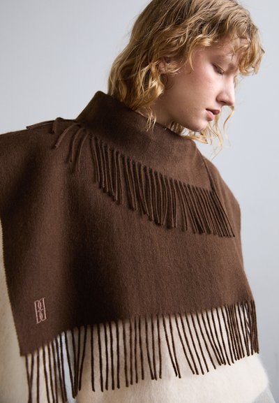 Poncho en laine marron avec un col montant, présentant de longues franges à l'ourlet et une section crème plus claire en bas. Détail de logo subtil sur le tissu.