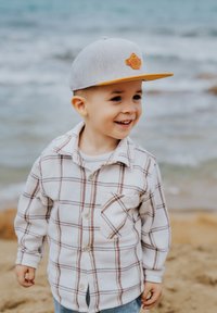 Chemise en flanelle blanche à carreaux beiges et marron, avec une poche sur la poitrine. Casquette grise avec visière orange et logo. Océan en arrière-plan.