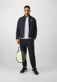 Marineblauwe zip-up jas met textuurdesign, wit T-shirt eronder, bijpassende broek en zwarte sneakers, met een tennisracket in de hand.