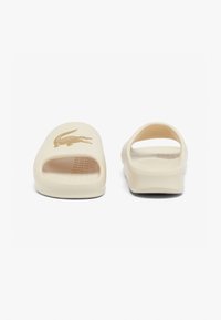 Beige slide sandals featuring a crocodile logo. Open-toe design with a textured footbed and smooth finish, suitable for casual wear.