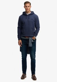 Sudadera con capucha azul marino con bolsillo frontal, forro interior gris y cordón ajustable. Se lleva con jeans oscuros ajustados y una camisa azul a cuadros anudada en la cintura.