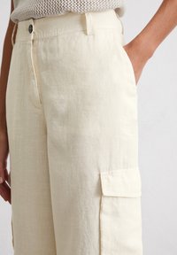 Helle beige Leinenhose mit entspannter Passform, ausgestattet mit seitlichen Taschen und einer einzelnen Cargotasche sowie einer strukturierten Oberfläche.