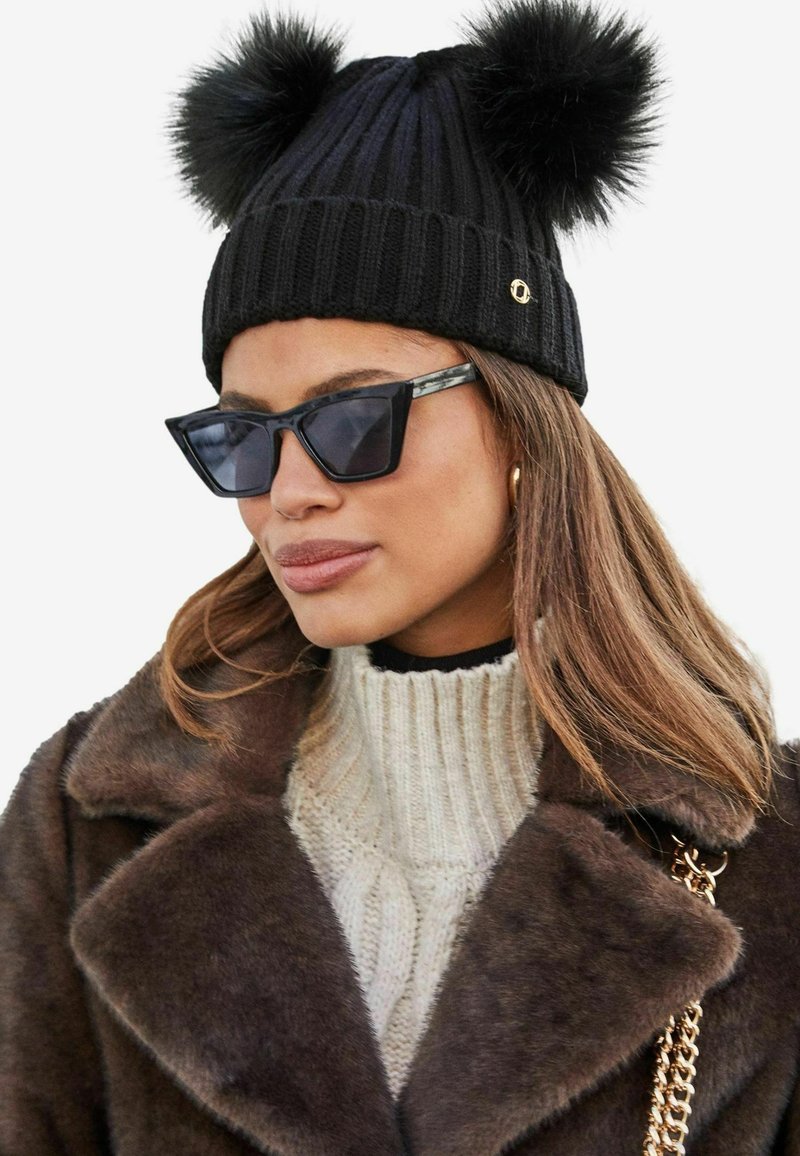 Gorro de punto negro con dos pompones de piel en la parte superior; textura acanalada; combinado con gafas de sol negras angulares y un suéter de cuello vuelto color crema.
