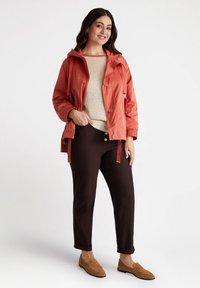 Giacca impermeabile leggera color corallo con cappuccio, chiusura a bottoni e lacci laterali. Abbinata a un top in maglia crema e pantaloni marroni scuri.