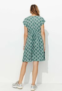Robe à manches courtes verte à motifs avec une taille cintrée et une jupe évasée. Le tissu a une finition texturée ; portée avec des baskets argentées.