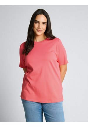 Kobieta o długich ciemnych włosach, ubrana w koralowo-różową koszulkę z krótkim rękawem i jasnoniebieskie dżinsy, stojąca na jednolitym jasnym tle.