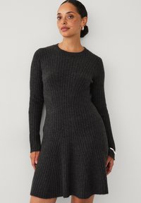 Frau mit Sommersprossen in einem dunkelgrauen, langärmeligen, gerippten Strickkleid, die vor einer schlichten hellen Wand steht.