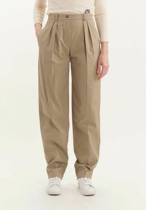 Pantalons plissés beiges à taille haute, avec poches latérales et fermeture à un bouton, faits d'un tissu lisse. Portés avec des baskets blanches.