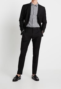 Blazer noir et pantalon associés à une chemise à carreaux noir et blanc. Le matériau semble lisse ; les chaussures sont en cuir noir poli.