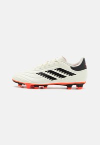Botín de fútbol blanco con logo de tres rayas negras, acentos rojos en la suela y parte superior texturizada. Presenta un diseño con cordones para un ajuste seguro.