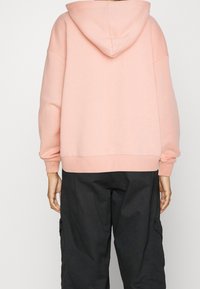 Felpa oversize rosa pastello con cappuccio foderato, polsini e orlo a costine e spalle scese, abbinata a pantaloni cargo neri.