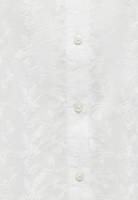 Tela blanca con sutiles patrones florales bordados y una fila vertical de tres pequeños botones blancos.