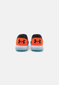 Baskets de sport noires et orange avec un dessus texturé, comportant une semelle intermédiaire blanche et une semelle extérieure bleue, arborant le logo Under Armour.
