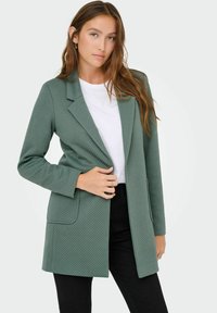 Femme portant un blazer texturé vert sur un haut blanc et un pantalon noir, tenant le blazer d'une main.