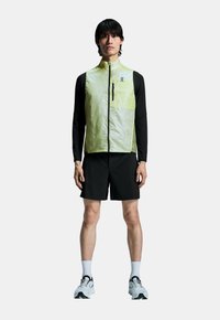 Lette gule vest med lynlås, parret med en sort langærmet skjorte og sorte shorts. Har en lomme på brystet.