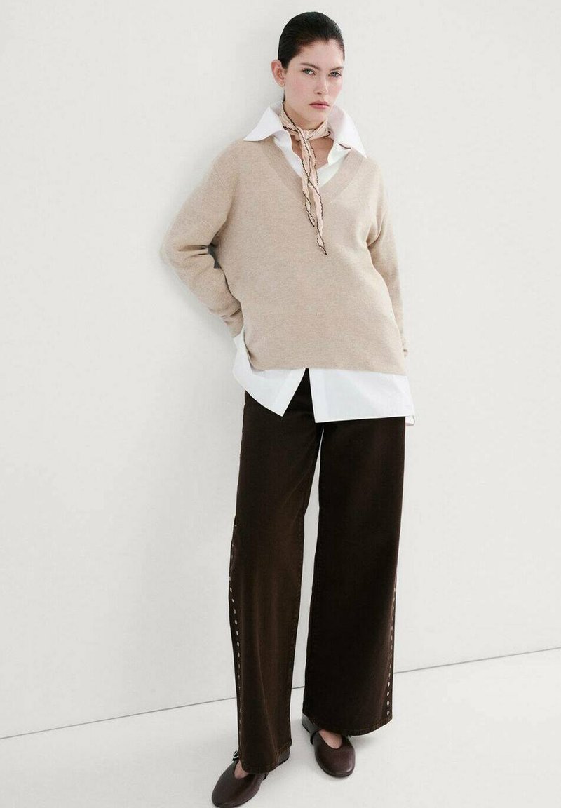 Kvinde iført beige V-hals sweater over hvid skjorte, brune bukser med vide ben og knapdetaljer i siden, mørkebrune sko og et lyst tørklæde omkring halsen.