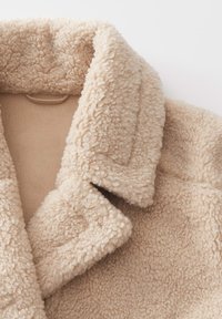 Manteau en fausse fourrure beige avec un large col à revers, texture douce et doublure intérieure lisse. Détails comprenant une boucle de suspension au niveau du cou.