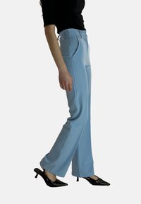 Lichtblauwe, op maat gemaakte broek met een hoge taille, wijde pijpen en plooien aan de voorkant, gecombineerd met zwarte puntschoenen met hak. Gladde stoftextuur.