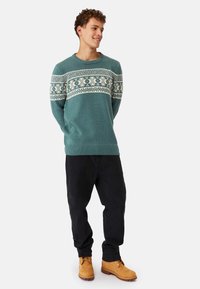 Teal knitted sweater with white Fair Isle pattern across chest, paired with black pants and brown boots. Designed with a round neckline and ribbed cuffs.