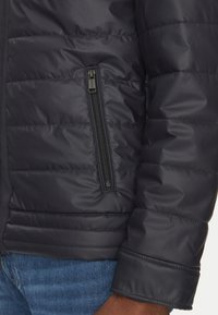 Chaqueta acolchada negra con un bolsillo con cremallera en el lado izquierdo. Presenta una textura acolchada y un ajuste ceñido en los puños.
