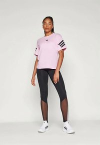 Ružová tričko s krátkym rukávom a čiernymi trojpruhovými akcentmi, skombinované s čiernymi legínami s sieťovanými panelmi a bielymi teniskami s čiernymi pruhmi.