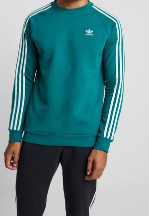 Mand iført teal Adidas-sweatshirt med hvide striber på ærmerne og logo på brystet, kombineret med sorte bukser med hvide striber.