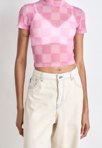 Růžový kostkovaný síťovaný crop top s vysokým límcem a krátkými rukávy, doplněný smetanovými džíny s vysokým pasem a modrým prošíváním.