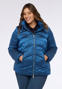 Chaqueta acolchada azul con cremallera frontal, patrón de chevron, cuello alto y bolsillos laterales. El material tiene una textura suave y brillante.