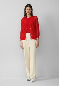 Rote offenmaschige Strickjacke mit V-Ausschnitt, Knopfverschluss und langen Ärmeln, über einem roten enganliegenden Oberteil getragen. Kombiniert mit cremefarbenen, weiten Hosen.