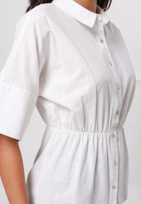 Camicia bianca a maniche corte realizzata in tessuto leggero, con un apertura frontale a bottoni e una vita arricciata, caratterizzata da un leggero motivo testurizzato.