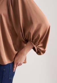 Brun, let bluse med voluminøse, samlede ærmer. Glat stoftekstur og elastiske manchetter. Parret med blå denim.