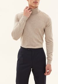 Beige turtleneck sweater i blødt stof, tætsiddende design, lange ærmer; båret med mørkeblå skræddersyede bukser.