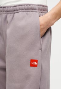 Χέρι ατόμου στην τσέπη ανοιχτόμωβ φόρμας με ελαστική μέση και κόκκινο εμβληματικό σημείο The North Face στο μηρό.