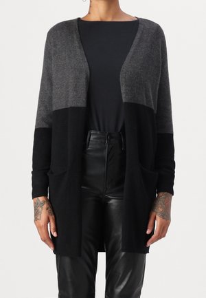 Henkilö, joka pukeutunut harmaaseen ja mustaan avoimeen neuletakkiin mustan paidan ja mustien nahkahousujen päällä, näyttää kukkatatuointeja kummassakin kädessä.