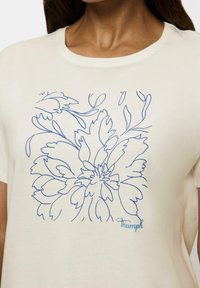 Kremowa bawełniana koszulka z niebieskim, rysowaną linią kwiatowym wzorem oraz słowem "Triumph" w skrypcie w prawym dolnym rogu.