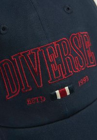 Granatowy daszek z czerwoną haftowaną napisami "DIVERSE" i "ESTD 1993." Posiada mały akcent w postaci trójkolorowej flagi pod tekstem, wykonany z materiału bawełnianego.