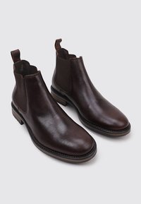 Botas de cuero marrón hasta el tobillo con paneles laterales elásticos, punta redondeada y suelas en contraste. Presentan una textura suave con costuras mínimas.